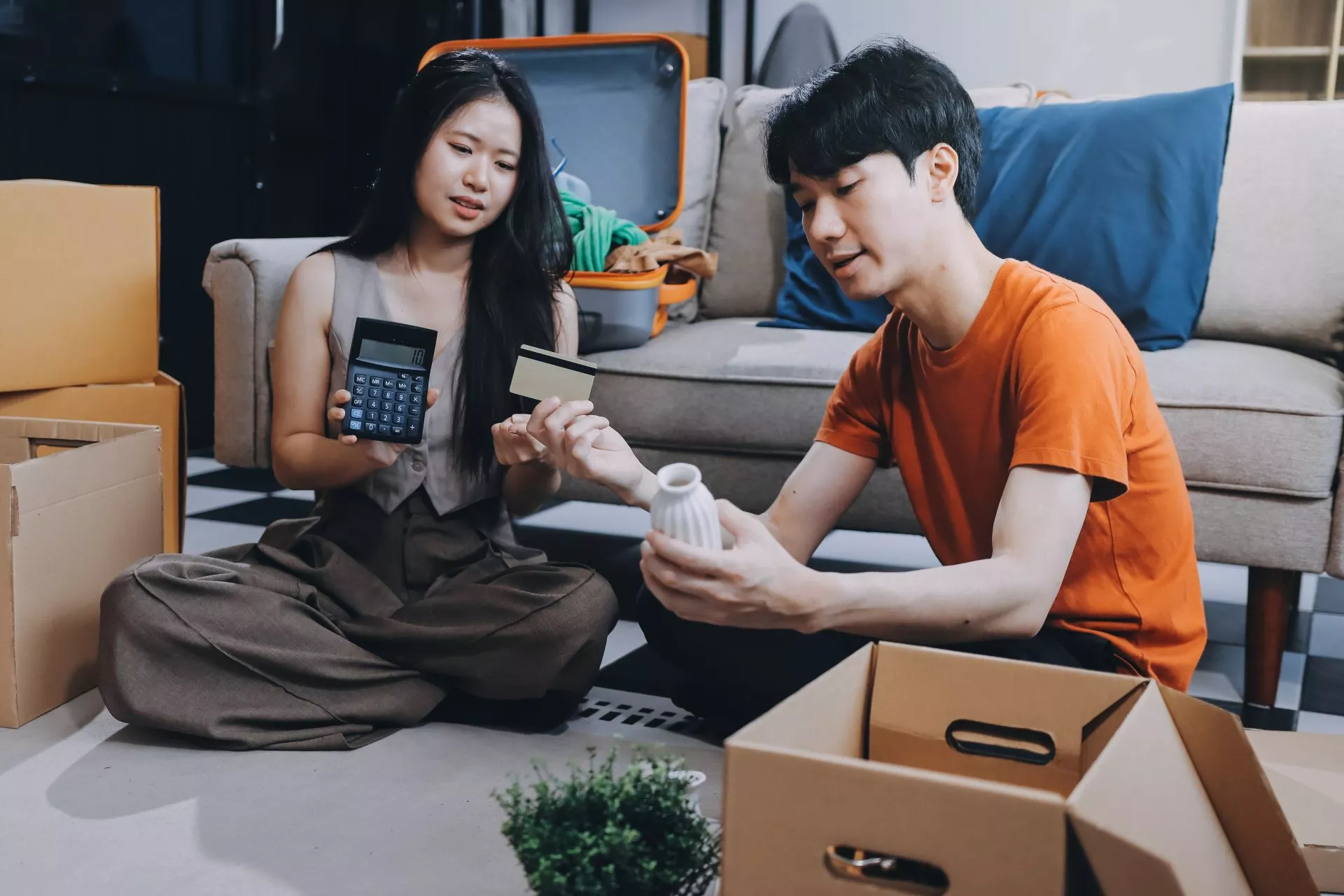 一對年輕情侶坐在地板整理搬家紙箱，女子手持計算機與信用卡，男子檢視家用品並討論開銷規劃的居家情境。