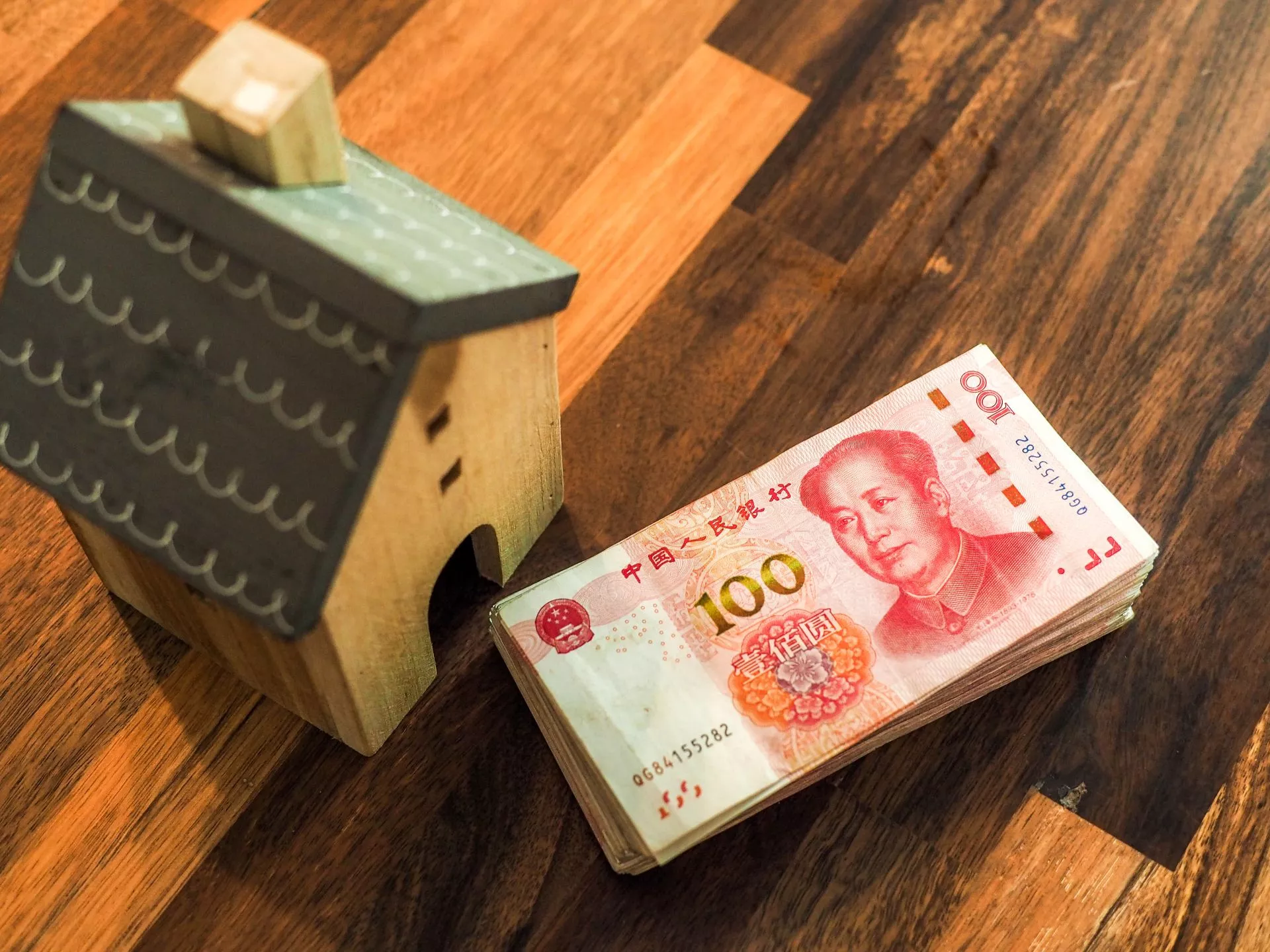 木質小屋模型旁放著一疊人民幣百元鈔票，置於木質桌面上，象徵房產與資金或購屋財務概念。