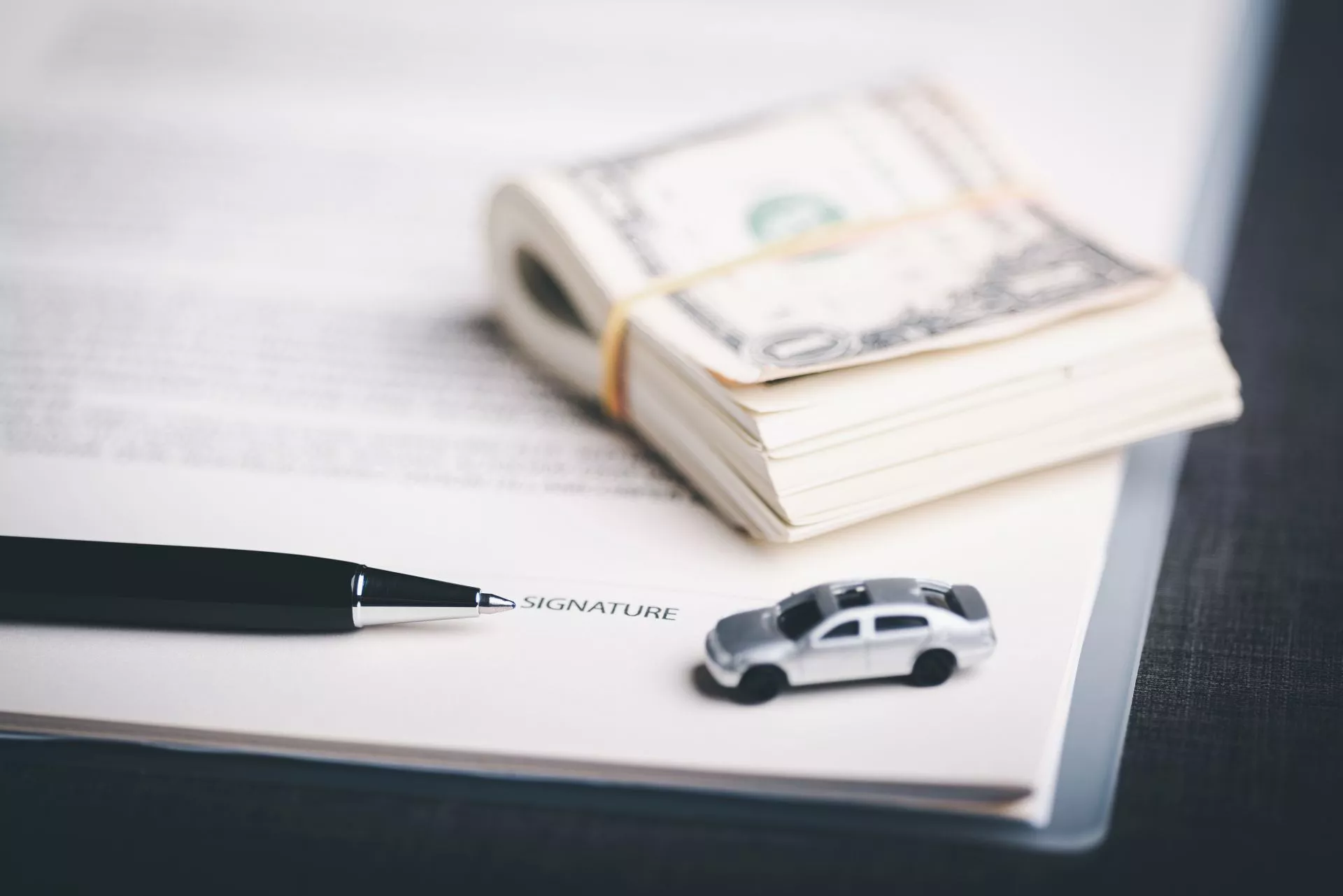 桌面上放著一疊綁著橡皮筋的現金、簽名文件與筆，旁邊有一台小型汽車模型，象徵汽車借款、資金周轉與貸款合約簽署情境。